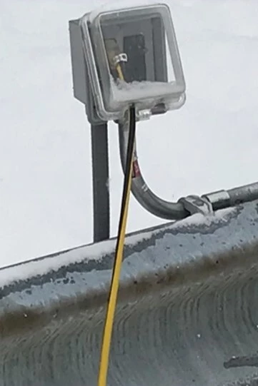 An outdoor Ground Fault Interrupter (GFI) outlet has a black and yellow electric cable plugged into it during the winter.